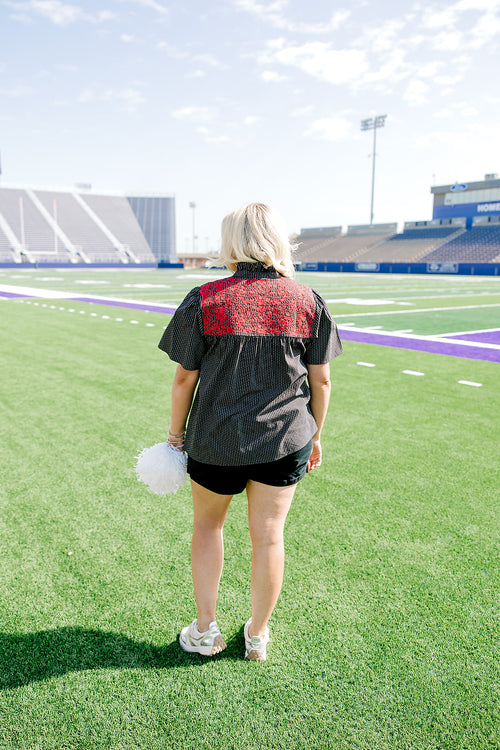Woman with blonde hair standing in a back profile position wearing the Reece Top in a lifestyle scene. The focal point of this image is the red floral embroidery on back of top as well as pleating on back. The model paired this top with black denim shorts. 