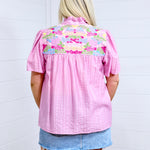 Woman with blonde hair standing in a back profile position wearing the Reece against a white background. The focal point of this image is the embroidery across the shoulders and pleating down the back of the top. The model paired this top with a light-wash denim skirt.