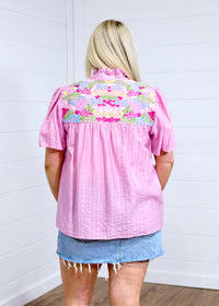 Woman with blonde hair standing in a back profile position wearing the Reece against a white background. The focal point of this image is the embroidery across the shoulders and pleating down the back of the top. The model paired this top with a light-wash denim skirt.