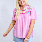 Woman with blonde hair standing in a front profile position wearing the Reece Top against a white background. The focal point of this image is the details on front of top as well as the intricate fabric detail. The model paired this top with a light-wash denim skirt. 