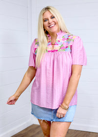 Woman with blonde hair standing in a front profile position wearing the Reece Top against a white background. The focal point of this image is the details on front of top as well as the intricate fabric detail. The model paired this top with a light-wash denim skirt. 