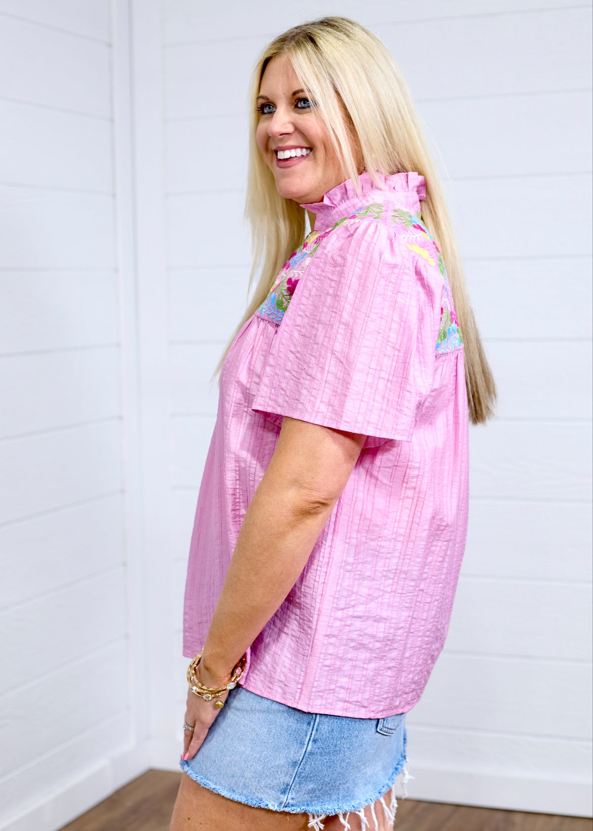 Woman with blonde hair standing in a side profile position wearing the Reece against a white background. The focal point of this image is the pleating down the sleeve as well as the high ruffled collar. The model paired a light-wash denim skirt with this top. 
