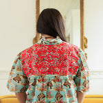 Woman with dark hair standing in a back profile position wearing the Reece Top in a lifestyle scene. The focal point of this image is the buckin' horse graphic as well as red embroidery across back. 