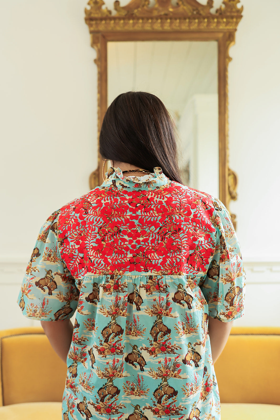 Woman with dark hair standing in a back profile position wearing the Reece Top in a lifestyle scene. The focal point of this image is the buckin' horse graphic as well as red embroidery across back. 