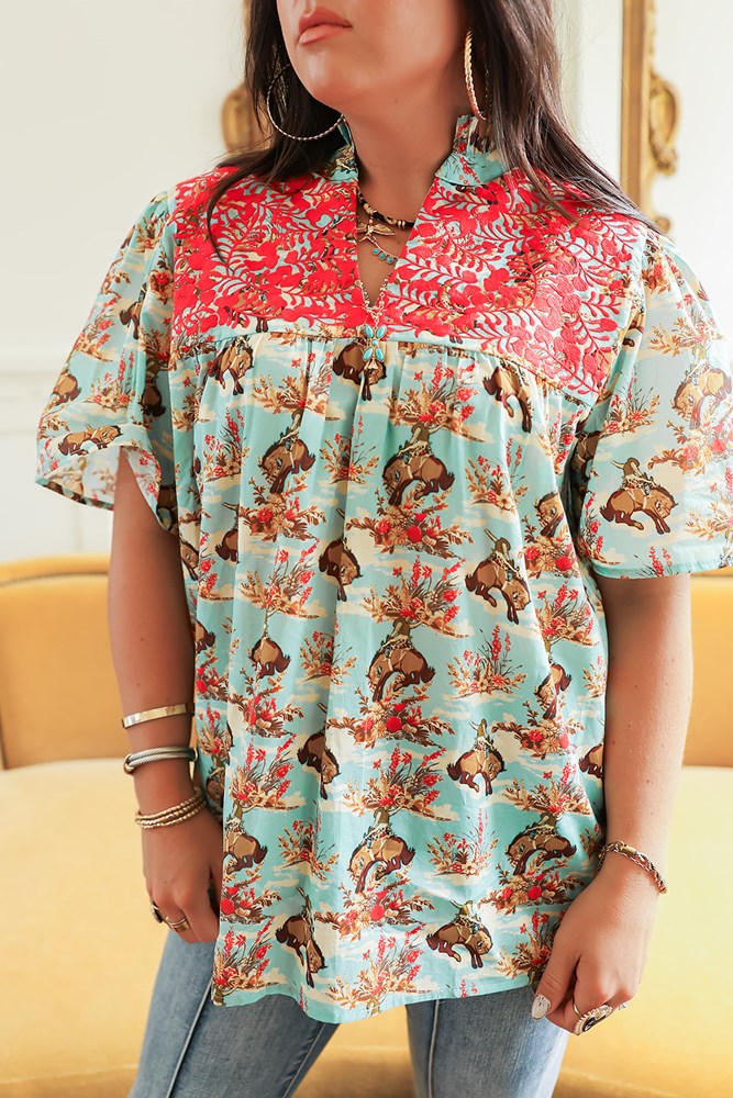 Woman with dark hair standing in a front profile position wearing the Reece Top in a lifestyle scene. The focal point of this image is the buckin horse graphic, red embroidery across chest, and v-neckline. The model paired this top with dark-wash denim jeans. 
