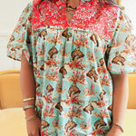 Woman with dark hair standing in a front profile position wearing the Reece Top in a lifestyle scene. The focal point of this image is the buckin horse graphic, red embroidery across chest, and v-neckline. The model paired this top with dark-wash denim jeans. 
