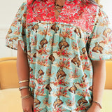 Woman with dark hair standing in a front profile position wearing the Reece Top in a lifestyle scene. The focal point of this image is the buckin horse graphic, red embroidery across chest, and v-neckline. The model paired this top with dark-wash denim jeans. 