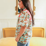 Woman with dark hair standing in a side profile position wearing the Reece Top in a lifestyle scene. The focal point of this image is the buckin' horse graphic as well as the red embroidery across shoulders. The model paired this top with dark-wash denim jeans. 