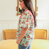 Woman with dark hair standing in a side profile position wearing the Reece Top in a lifestyle scene. The focal point of this image is the buckin' horse graphic as well as the red embroidery across shoulders. The model paired this top with dark-wash denim jeans. 