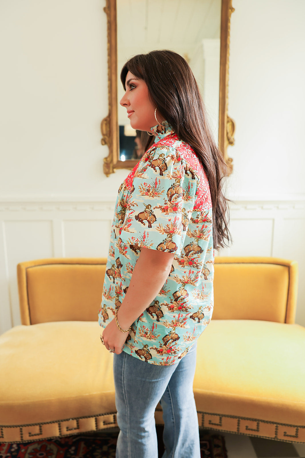 Woman with dark hair standing in a side profile position wearing the Reece Top in a lifestyle scene. The focal point of this image is the buckin' horse graphic as well as the red embroidery across shoulders. The model paired this top with dark-wash denim jeans. 