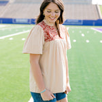 Woman standing to the side with a short-sleeve orange top featuring burnt orange embroidery, and dark-wash denim shorts. 