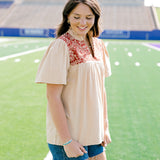 Woman standing to the side with a short-sleeve orange top featuring burnt orange embroidery, and dark-wash denim shorts. 