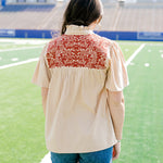 Woman with dark hair standing in a back profile position wearing a short-sleeve orange top with burnt orange embroidery, and dark-wash denims shorts. 