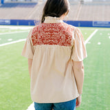 Woman with dark hair standing in a back profile position wearing a short-sleeve orange top with burnt orange embroidery, and dark-wash denims shorts. 