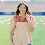 Woman facing forward wearing a short-sleeve orange shirt with burnt orange embroidery and dark-wash denim shorts. 