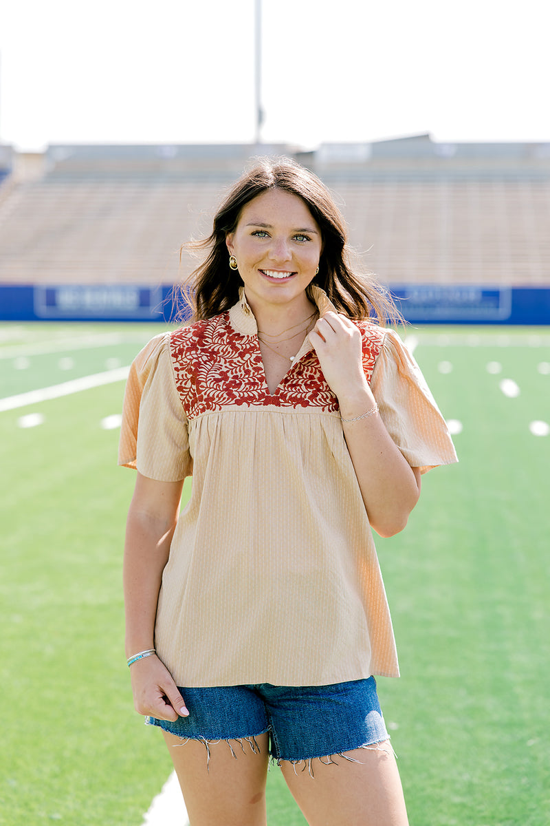 Woman facing forward wearing a short-sleeve orange shirt with burnt orange embroidery and dark-wash denim shorts. 