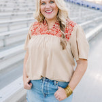 Woman with blonde hair facing forward with a short-sleeve orange top with burnt orange embroidery, and a light-wash denim skirt. 