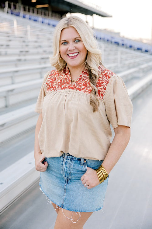 Woman with blonde hair facing forward with a short-sleeve orange top with burnt orange embroidery, and a light-wash denim skirt. 