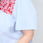 Woman with dark hair standing at a side profile position in the Reece Top against a white background. The focal point of this image is in a deep focal length to show stripe detailed fabric and red embroidery across shoulders. 