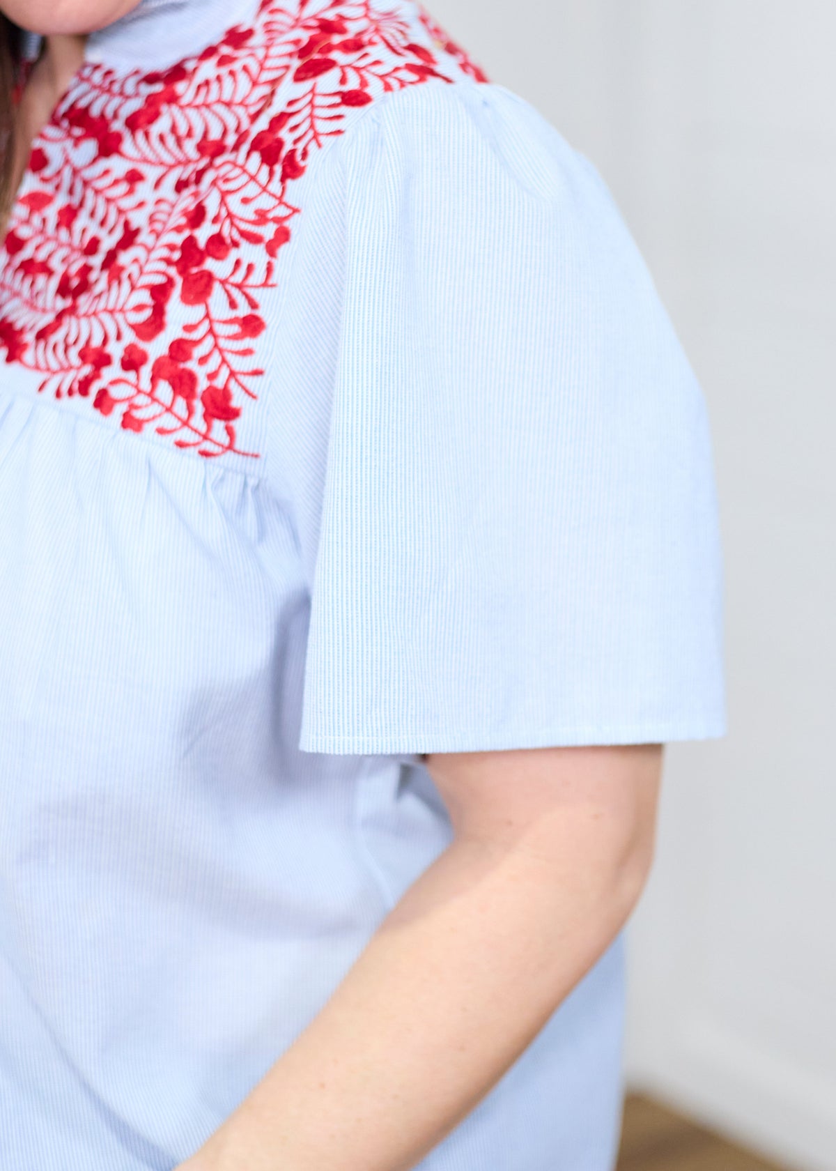 Woman with dark hair standing at a side profile position in the Reece Top against a white background. The focal point of this image is in a deep focal length to show stripe detailed fabric and red embroidery across shoulders. 