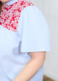Woman with dark hair standing at a side profile position in the Reece Top against a white background. The focal point of this image is in a deep focal length to show stripe detailed fabric and red embroidery across shoulders. 