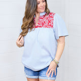 Woman with dark hair standing in a front profile position wearing the Reece Top against a white background. The focal point of this image is the red embroidery across the chest and v-neckline. The model paired this top with dark-wash denim shorts.