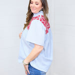 Woman with dark hair standing in a side profile position wearing the Reece Top against a white background. The focal point in this image is the sleeve length, shirt length and embroidery across shoulders. The model paired this top with dark-wash denim shorts. 