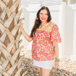Woman with dark hair is standing in a front profile position wearing the Reece Top in a lifestyle background. The focal point of this image is the top in its entirety as well as the "real time" scenery. The model has paired this top with a white skirt. 
