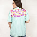 Woman with dark hair standing in a back profile position wearing the Reece Top against a white background. The focal point of this image is the embroidery on the shoulders of the top. The model paired this top with dark-wash denim shorts. 