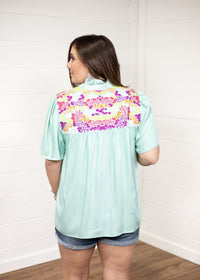 Woman with dark hair standing in a back profile position wearing the Reece Top against a white background. The focal point of this image is the embroidery on the shoulders of the top. The model paired this top with dark-wash denim shorts. 