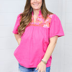 Woman with dark hair standing in front profile position wearing the Reece Top against a white background. The focal point of this image is the embroidery on chest, pleating beneath, and v-neckline. The model paired this top with dark-wash denim shorts.