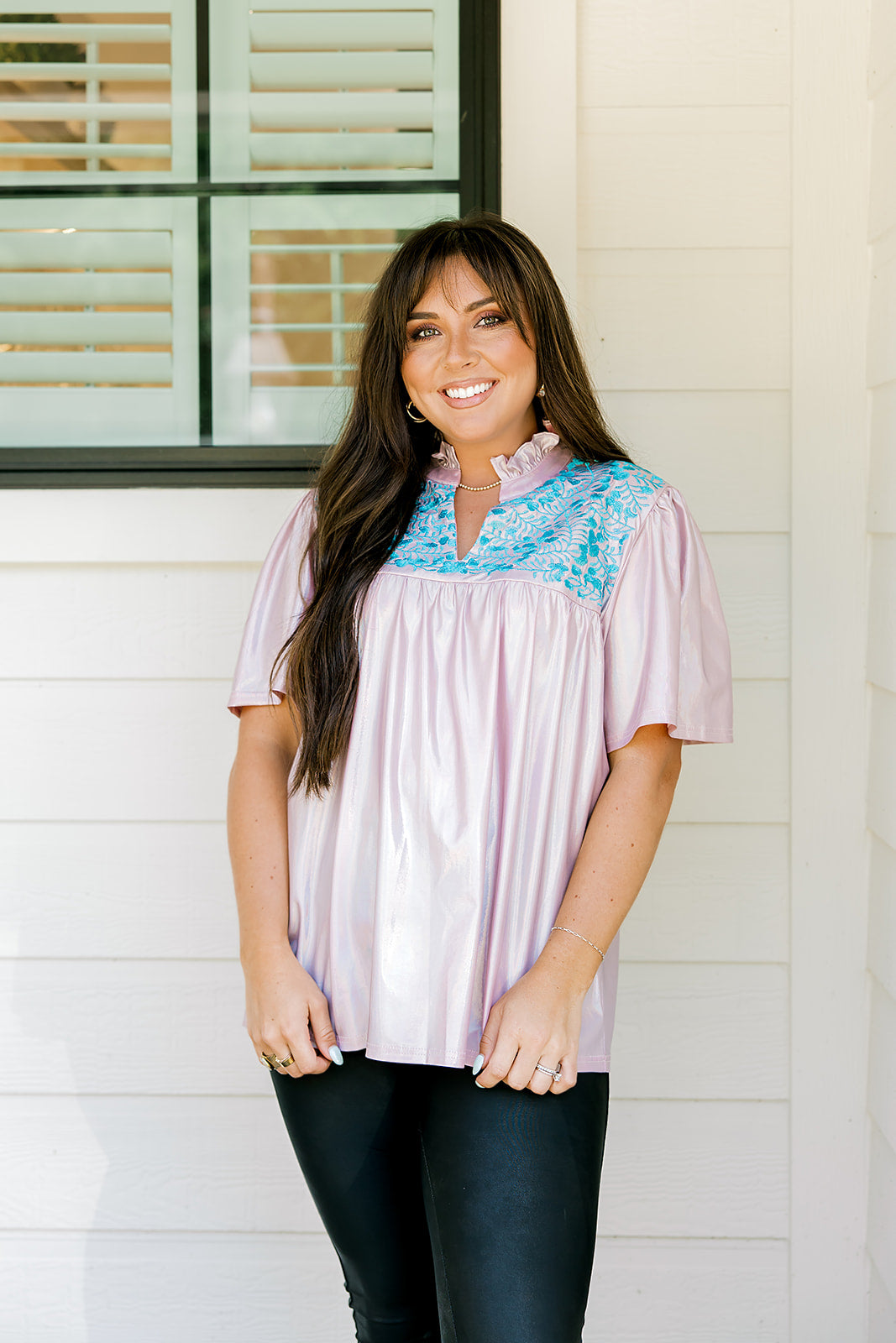 Woman with dark hair wearing a top in the Reece body style. Fabric is pink iridescent and is accented with turquoise embroidery along shoulders and chest. Model paired this top with faux leather leggings. 