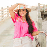 Woman with dark hair standing in a front facing position wearing the Reece Top in a lifestyle scene. The focal point of this image is the bright pink shirt with pleating underneath embroidery. The model paired this top with white denim shorts.