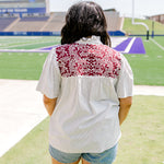 Woman with dark hair standing in a back profile position wearing the Reece Top in a lifestyle scene. The focal point of this image is the maroon embroidery on the back of the top as well as ruffled collar and striped graphics along shirt. The model paired this top with light-wash denim shorts. 