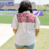 Woman with dark hair standing in a back profile position wearing the Reece Top in a lifestyle scene. The focal point of this image is the maroon embroidery on the back of the top as well as ruffled collar and striped graphics along shirt. The model paired this top with light-wash denim shorts. 