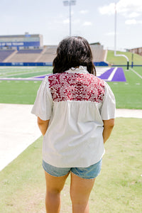 Woman with dark hair standing in a back profile position wearing the Reece Top in a lifestyle scene. The focal point of this image is the maroon embroidery on the back of the top as well as ruffled collar and striped graphics along shirt. The model paired this top with light-wash denim shorts. 