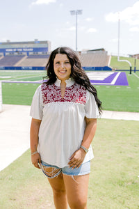 Woman with dark hair standing in a front profile position wearing the Reece Top in a lifestyle scene. The focal point of this image is the ruffled collar, v-neckline, and pleating on front of top. The model paired this top with light-wash denim shorts. 