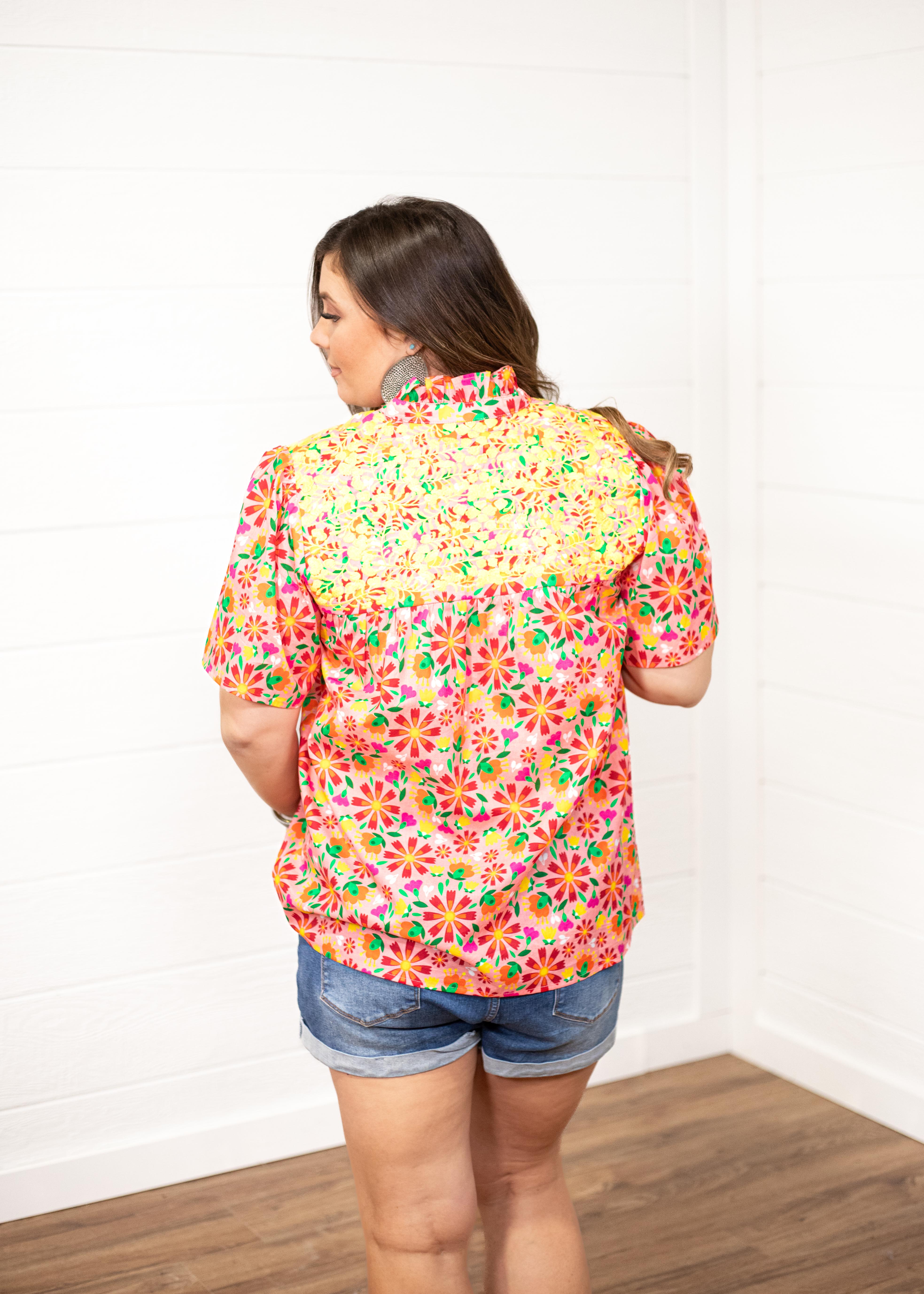 Woman with dark hair standing in a back profile position wearing the Reece Top against a white background. The focal point of this image is the embroidery across the shoulders as well as the graphics along the back of the top. The model has this top paired with dark-wash denim shorts.
