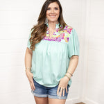 Woman with dark hair standing in a front profile position wearing the Reece Top standing against a white background. The focal point of this image is the embroidery across the chest of the top and the v-neckline. The model paired this top with dark-wash denim shorts. 