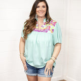 Woman with dark hair standing in a front profile position wearing the Reece Top standing against a white background. The focal point of this image is the embroidery across the chest of the top and the v-neckline. The model paired this top with dark-wash denim shorts. 