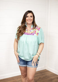 Woman with dark hair standing in a front profile position wearing the Reece Top standing against a white background. The focal point of this image is the embroidery across the chest of the top and the v-neckline. The model paired this top with dark-wash denim shorts. 