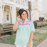 Woman with dark hair standing in a front profile position wearing the Reece Top standing in a lifestyle positioned background. The emphasis of this image is the model wearing the top in "real time" with emphasis on the embroidery, sleeves, and front of top. The model paired this top with white pants. 