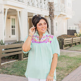 Woman with dark hair standing in a front profile position wearing the Reece Top standing in a lifestyle positioned background. The emphasis of this image is the model wearing the top in "real time" with emphasis on the embroidery, sleeves, and front of top. The model paired this top with white pants. 