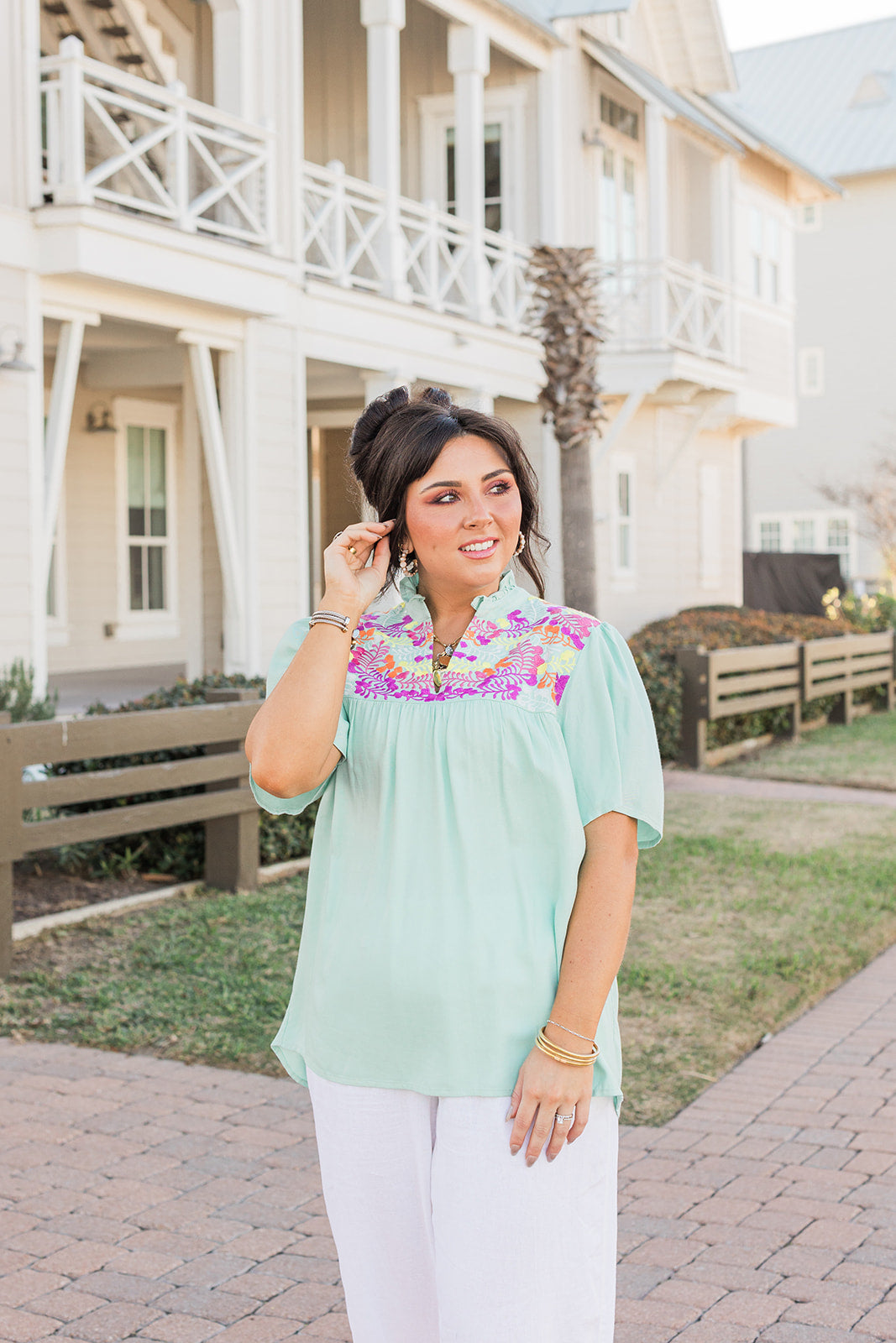 Woman with dark hair standing in a front profile position wearing the Reece Top standing in a lifestyle positioned background. The emphasis of this image is the model wearing the top in "real time" with emphasis on the embroidery, sleeves, and front of top. The model paired this top with white pants. 