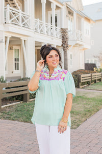 Woman with dark hair standing in a front profile position wearing the Reece Top standing in a lifestyle positioned background. The emphasis of this image is the model wearing the top in "real time" with emphasis on the embroidery, sleeves, and front of top. The model paired this top with white pants. 