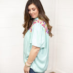 Woman with dark hair standing in a side profile position wearing the Reece Top standing against a white background. The focal point of this image is the ruffled sleeves and embroidery on top of the shoulder. The model paired this top with dark-wash denim shorts.