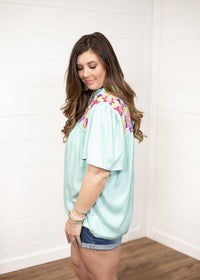 Woman with dark hair standing in a side profile position wearing the Reece Top standing against a white background. The focal point of this image is the ruffled sleeves and embroidery on top of the shoulder. The model paired this top with dark-wash denim shorts.