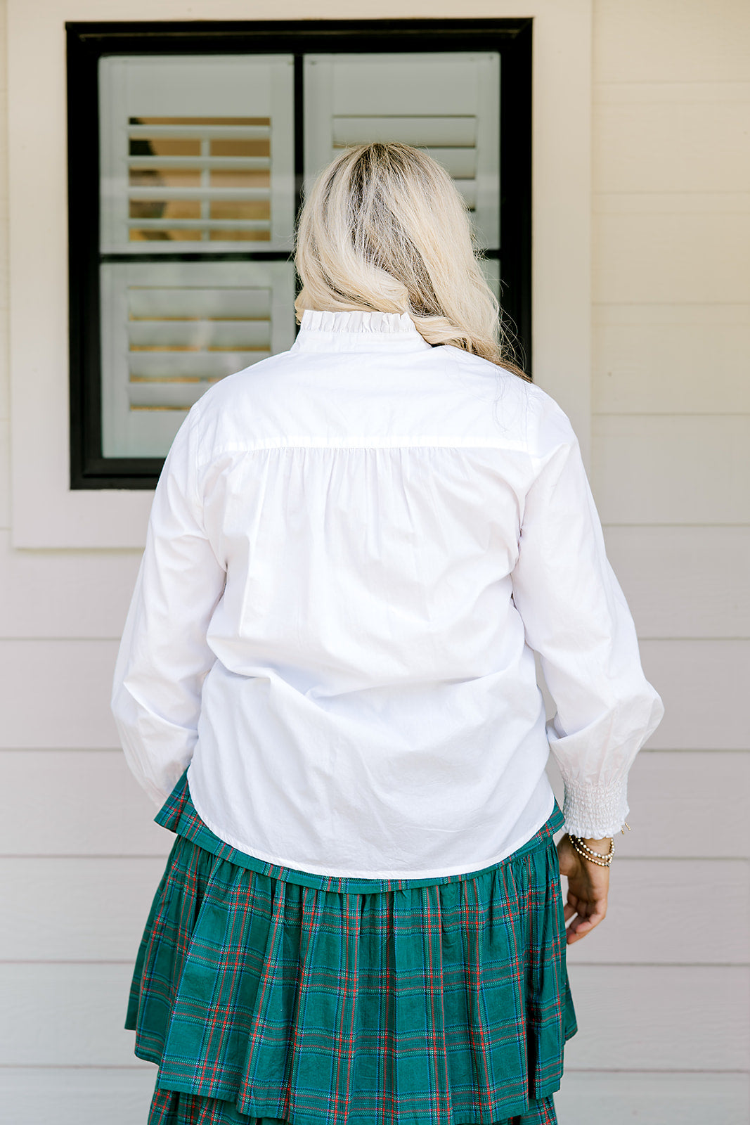 Woman with blonde hair wearing a white long-sleeve top with red ruby buttons down the front. Model paired this top with the Riley Ruffle Skirt. 