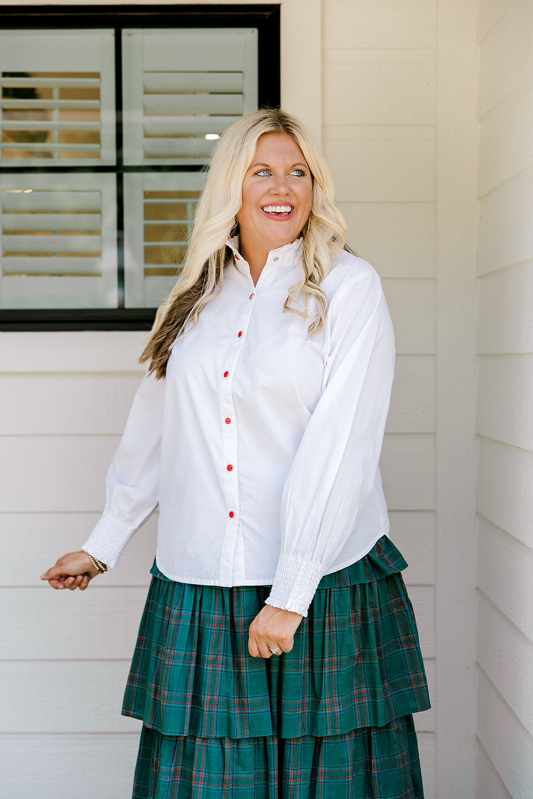 Woman with blonde hair wearing a white long-sleeve top with red ruby buttons down the front. Model paired this top with the Riley Ruffle Skirt. 