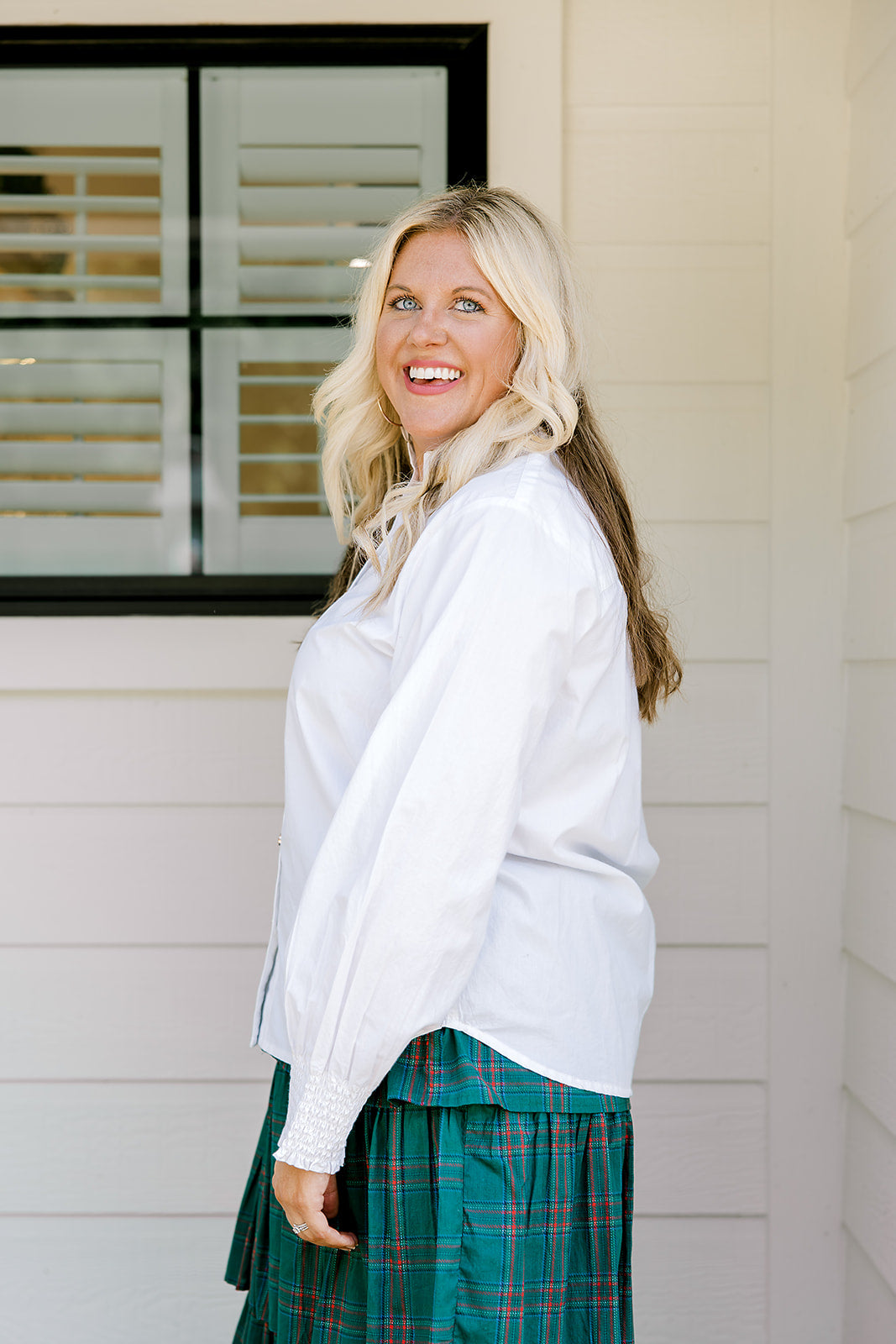 Woman with blonde hair wearing a white long-sleeve top with red ruby buttons down the front. Model paired this top with the Riley Ruffle Skirt. 